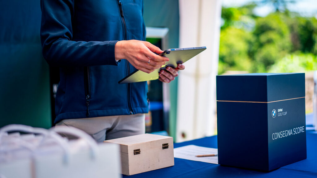 Una persona tiene in mano un tablet accanto a una scatola blu con la scritta "BMW Golf Cup" e "Consegna Score"