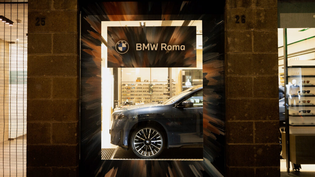 Vetrina con un'auto in mostra e il logo BMW Roma, incorniciati da una grafica astratta.