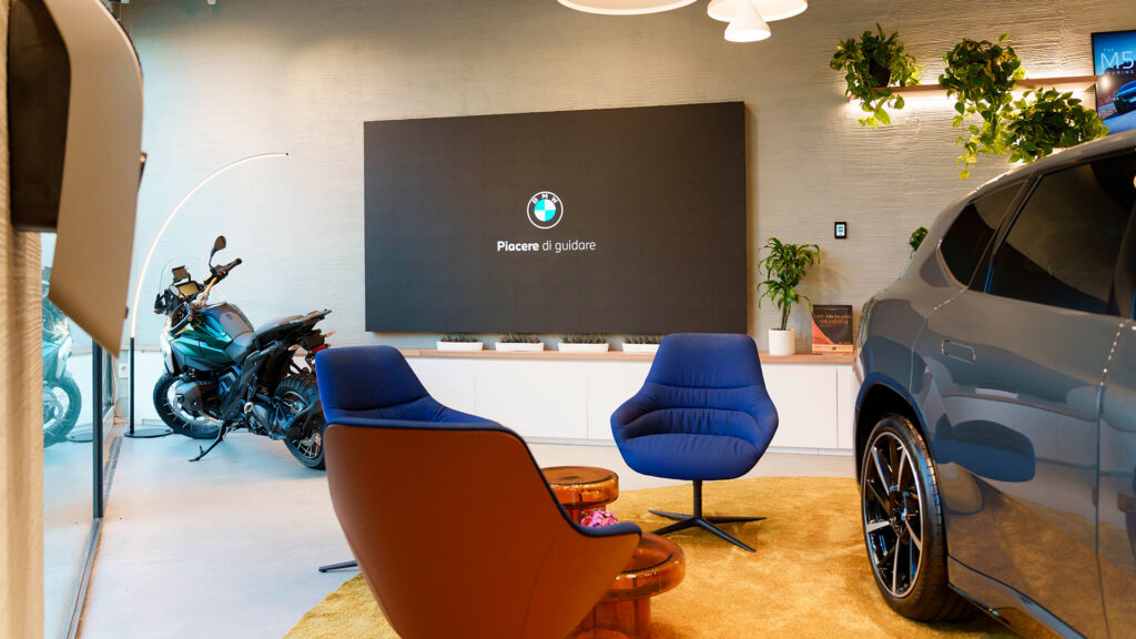 Showroom BMW con poltrone blu, una moto, un'auto e un grande schermo a parete.